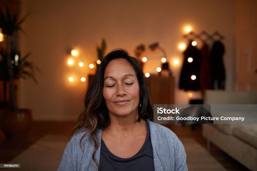 Serene mature woman smiling while meditating at home Serene woman smiling with her eyes closed while sitting on her living room floor practicing yoga Meditating Stock Photo