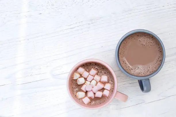 Hot chocolate in two mugs, one with small marshmallows, isolated on white painted wood from above. Space for text.