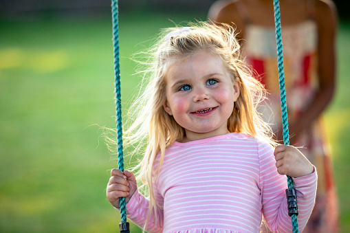 A front-view shot of a young girl playing on a swing in the garden, she is smiling and having fun.