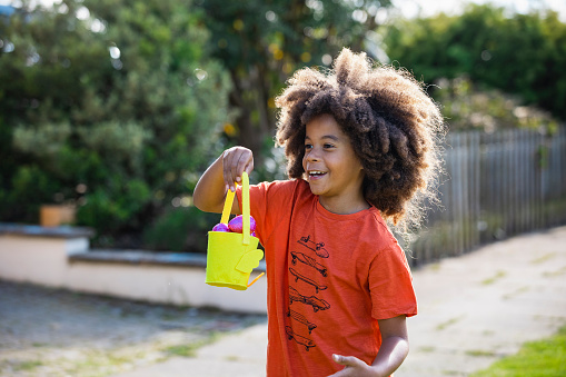A front-view portrait of a young boy with an afro, he is smiling while holding an Easter egg bucket.