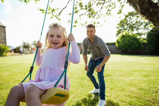 A close-up shot of a young girl playing on a swing in the garden, she is smiling with her father.