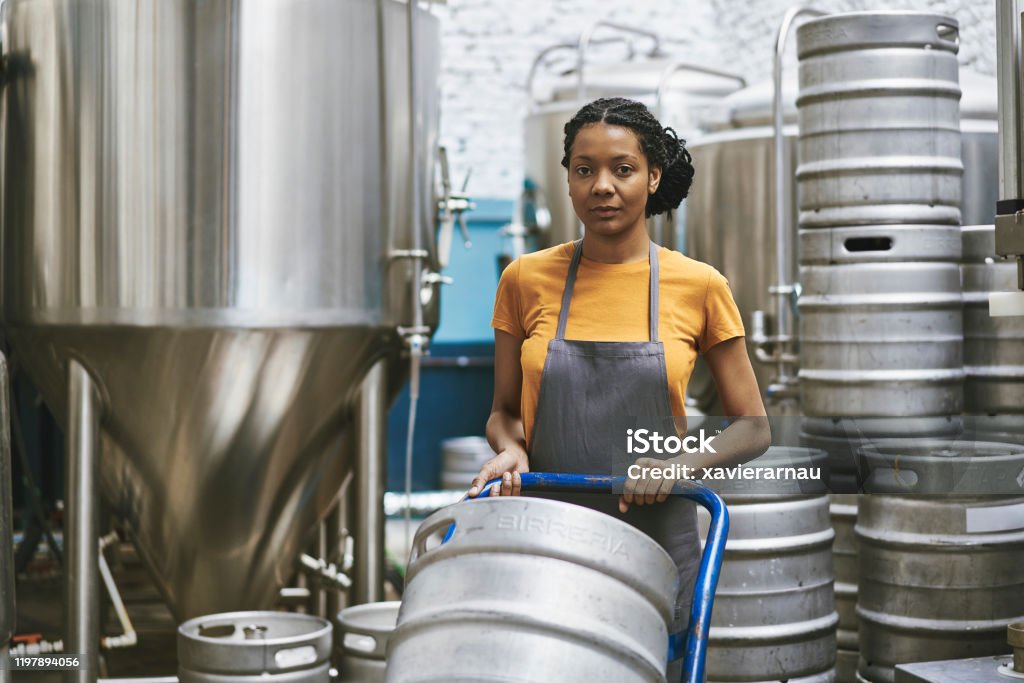 Mixed Race Female Craft Beer Brewer in Buenos Aires Hardworking mid adult female craft beer brewer looking at camera while pushing keg on hand truck in brewery interior. Hand Truck Stock Photo