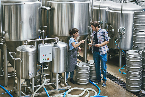 Elevated view of male and female Buenos Aires craft beer workers in their 30s standing among kegs and vats with digital tablet and conversing.