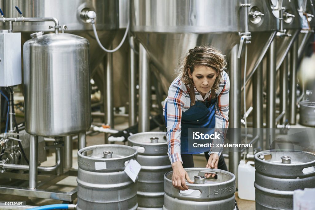 Hispanic Female Brewery Worker Lifting Beer Keg Close-up of female worker in mid 30s bending over to lift beer keg for washing at Buenos Aires craft brewery. Brewery Stock Photo