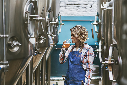 Female Buenos Aires craft brewer in mid 30s standing between vats and examining beer sample in glass.
