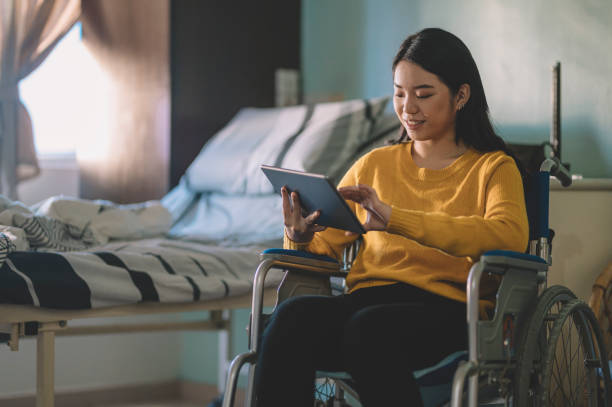 una adolescente asiática china en su silla de ruedas en la sala del hospital usando su tableta digital - patient room fotografías e imágenes de stock