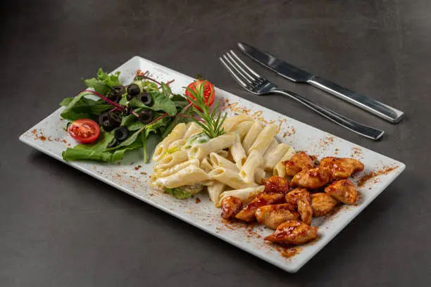 Grilled cubed chicken served with pasta and salad on white plate on dark stone table