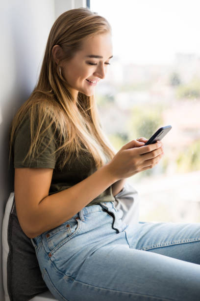 istock Gorgeous young woman checking email and sending sms message on mobile phone sitting on windowsill 1197905467