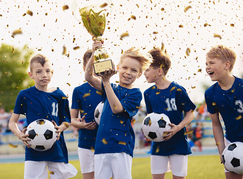 Happy kids in elementary school sports team celebrating soccer succes in tournament final game. Boys smiling and rising up golden cup on trophy ceremony