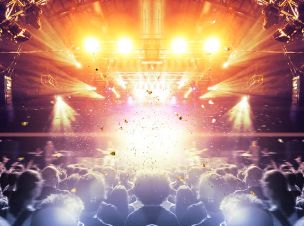 Concert stage, people are visible waving and clapping, silhouettes are visible