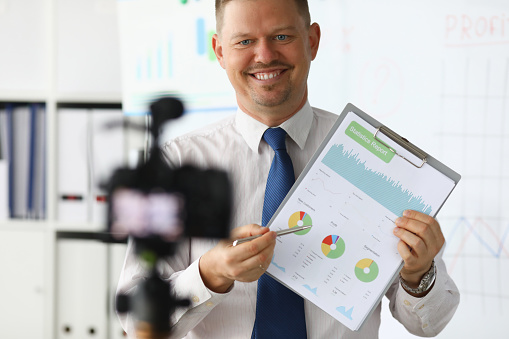 Portrait of smiling blogger in classic suit and tie showing statistics data with graphs charts diagram. Promo videoblog or photo session in office camcorder to tripod. Blurred background
