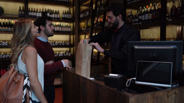 Happy couple saying goodbye to friendly sommelier packing a wine bottle in a paper bag at a wine cellar