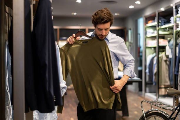 un tipo latinoamericano probando un suéter encima en una tienda de ropa para hombres - adicto a las compras fotografías e imágenes de stock