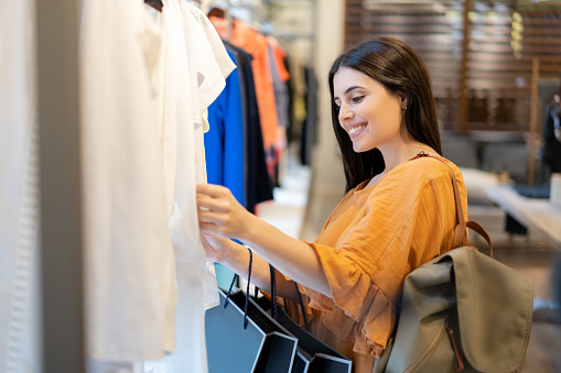 Side view of beautiful woman looking at different shirts on rack at a women's store - Consumerism concepts