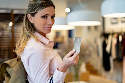 Young formal woman holding a credit card while purchasing clothes at a store smiling at camera - Consumerism concepts **DESIGN ON CREDIT CARD WAS MADE FROM SCRATCH BY US**