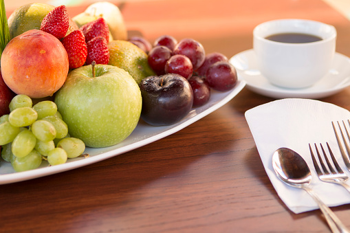 Plate of colorful various fruits with white cup of coffee and spoon and fork over a wood table with natural light