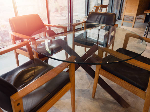 Modern round glass table with four wooden chairs in living room loft style near window glass with sunlight from outside.