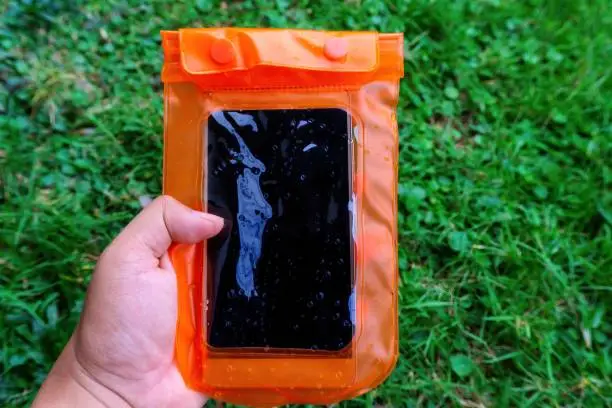 Woman hand holding the orange waterproof mobile phone case over the green grass background. PVC zip lock bag protect mobile phone and important items from water. Concept for Songkran water festival in Thailand.