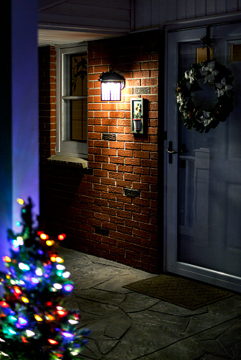Small, decorated, outdoor, holiday season tree with illuminated Christmas lights on the front porch at night welcoming all visitors - family and friends - to the warmly lit brick facade and decorated wreath front entrance door of a residential home.