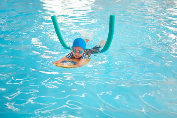 Cute little Asian toddler boy child kicking feet in swimming costume wear swimming goggles use pool noodle and kickboard learn to swim at indoor pool, Swimming school for small children concept