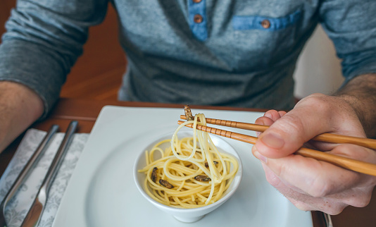 Unrecognizable man eating spaghetti and crickets with chopsticks