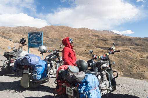 Solo Indian woman traveler trekker and Biker standing beside her motorcycle relaxing and looking at view after a long days hiking bike journey in high altitude Himalayan Mountain region, outing and exploring local hill and its culture. Adventure Tourism Photography. Kaza Himachal Pradesh India South Asia Pac October 2019