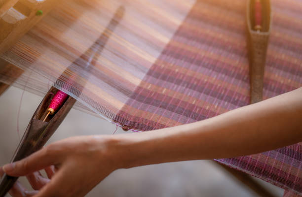 Woman working on weaving machine for weave handmade fabric. Textile weaving. Weaving using traditional hand weaving loom on cotton strands. Textile or cloth production in Thailand. Asian culture.