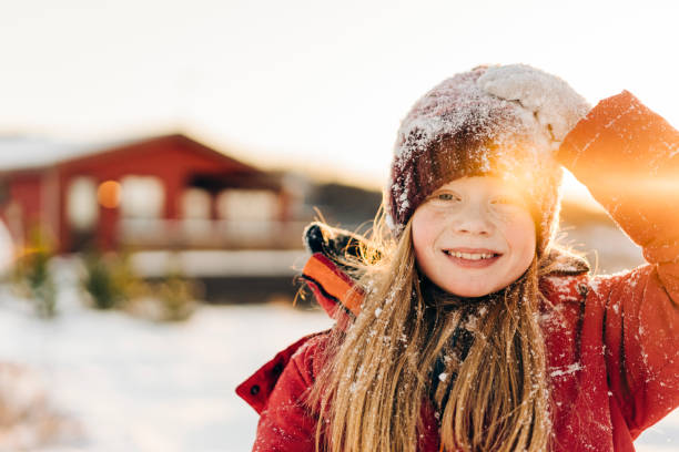 Close-up of smiling girl enjoying outdoors during winter. Portrait of cute blond female child is having snow on knit hat. She is wearing warm clothing against sky.