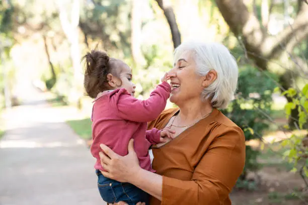 Senior grandmother holding her baby granddaughter in public park. Grandmother has white hair and is wearing an orange blouse. They are in outdoor. Selective focus on models. Shot with a full frame mirrorless camera.