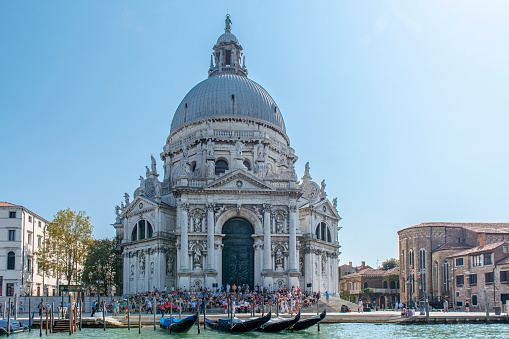 People sitting on the steps at the entrance of Santa Maria della Salute church (Saint Mary of Health) by the Grand Canal at Venice, Italy. The Grand Canal is an ancient waterway that goes from the Saint Mark Basin to a lagoon near the Santa Lucia rail station.\nThe city of Venice is an Unesco World Heritage site and it is visited daily by thousands of tourists from all over the world. Some gondolas are floating next to the sidewalk.