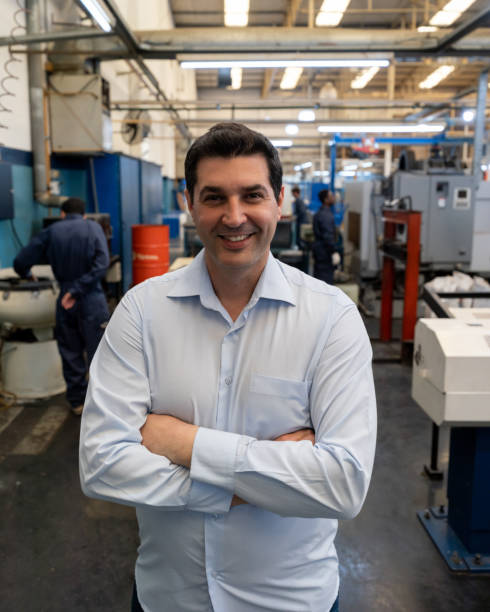 istock Cheerful engineer at a manufacturing plant looking at camera with arms crossed while blue collar workers work at background 1198042344