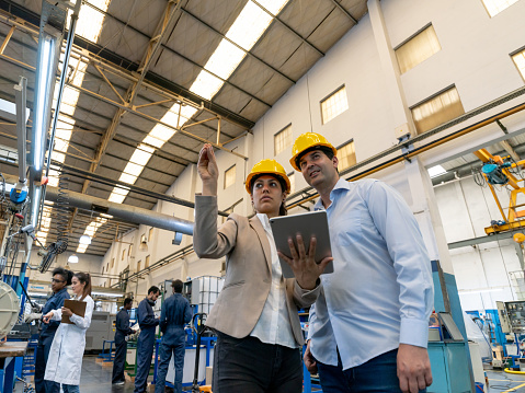 Female supervisor pointing away to male manager while she holds a tablet at a factory - Incidental people at background