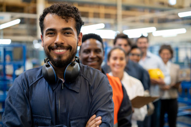 alegre apuesto trabajador de cuello azul y equipo de ingenieros en una fábrica de pie en una fila sonriendo a la cámara - manufacturing fotografías e imágenes de stock