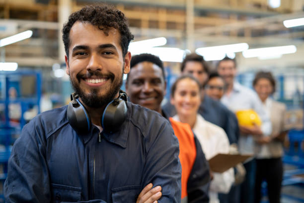 Cheerful handsome blue collar worker and team of engineers at a factory standing in a row smiling at camera Cheerful handsome blue collar worker and team of engineers at a factory standing in a row smiling at camera with arms crossed working class stock pictures, royalty-free photos & images