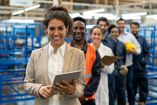 Hermosa gerente femenina en una fábrica que sostiene una tableta y un equipo de trabajadores de cuello azul, ingenieros e inspectores de pie en una fila sonriendo a la cámara photo