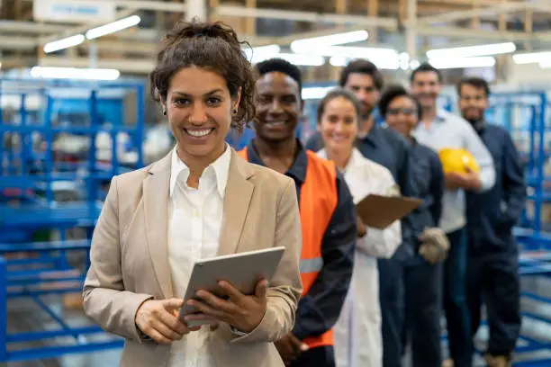 Beautiful female manager at a factory holding a tablet and team of blue collar workers, engineers and inspectors standing in a row smiling at camera very cheerfully