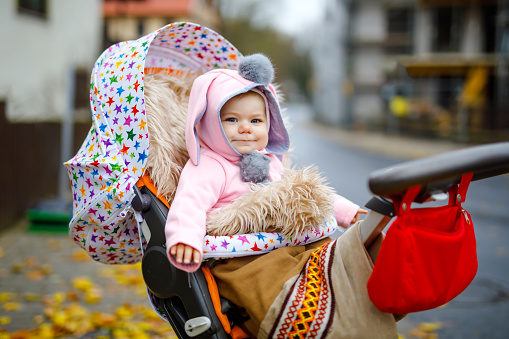 Cute little beautiful baby girl sitting in the pram or stroller on autumn day. Happy smiling child in warm clothes, fashion stylish pink baby coat with bunny ears. Baby going on a walk with parents