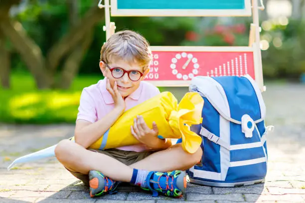 Happy little kid boy with glasses sitting by desk and backpack or satchel. Schoolkid with traditional German school bag cone called Schultuete on his first day to school.Back to school