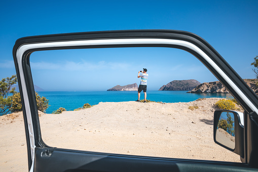 Man standing at the edge of a cliff and taking photos (Milos island, Cyclades, Greece). View from the car.