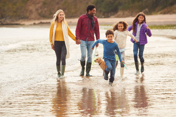 Multi-Cultural Family With Pet Dog Walking Along Beach Shoreline On Winter Vacation Multi-Cultural Family With Pet Dog Walking Along Beach Shoreline On Winter Vacation five people photos stock pictures, royalty-free photos & images
