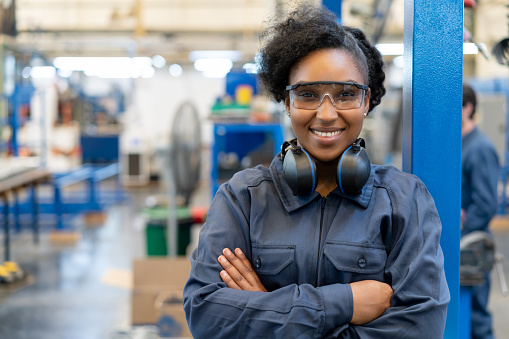 Confident black female engineer at a manufacturing plant facing camera smiling with arms crossed wearing protective gear