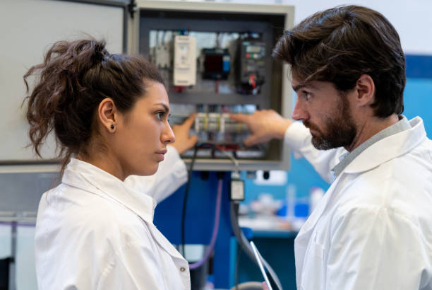 istock Male and female inspectors looking at each other while pointing at a switch board in a factory very serious 1198074866