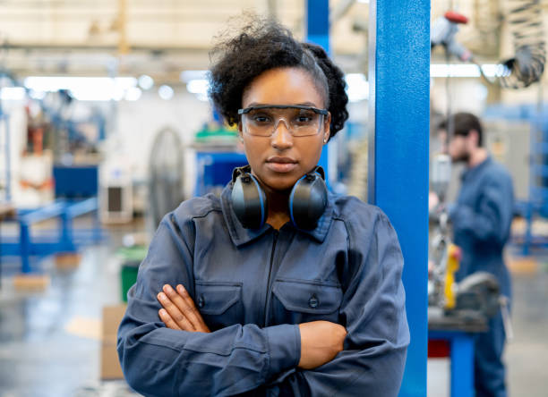 mujer joven negra y segura de sí misma que trabaja en una fábrica de fabricación con gafas protectoras mirando la cámara con los brazos cruzados - industry portrait production line factory fotografías e imágenes de stock