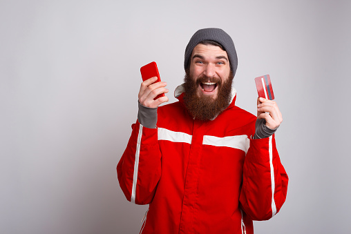 Amazed bearded hipster man wearing winter clothes and holding smartphone and credit card