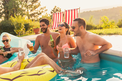 Group of friends at a poolside summer party, having fun in the swimming pool, drinking cocktails and beer and making a toast