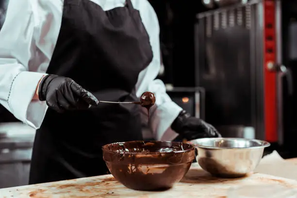 cropped view of chocolatier in black apron holding stick with tasty candy near chocolate in bowl