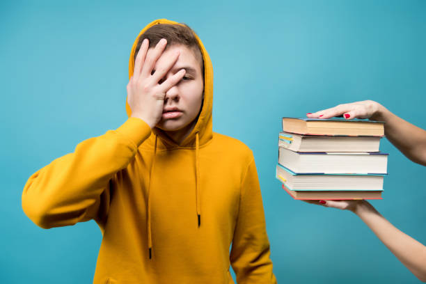 a teenager makes facepalm while he is offered a stack of books. guy expresses unwillingness to learn - unwillingness imagens e fotografias de stock