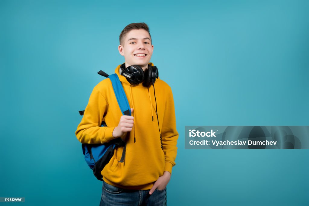 tipo con auriculares en una sudadera amarilla sobre un fondo azul con una mochila - Foto de stock de Chicos adolescentes libre de derechos