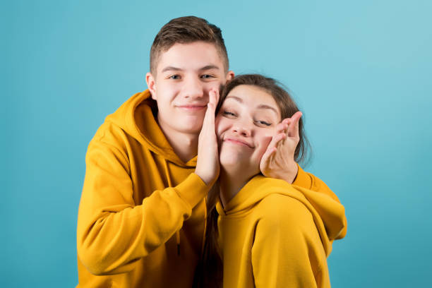 brother squeezes the cheeks of an older sister, fooling around on a blue background. brother squeezes the cheeks of an older sister, fooling around on a blue background. Close-up, teenagers in yellow hoodies brother stock pictures, royalty-free photos & images