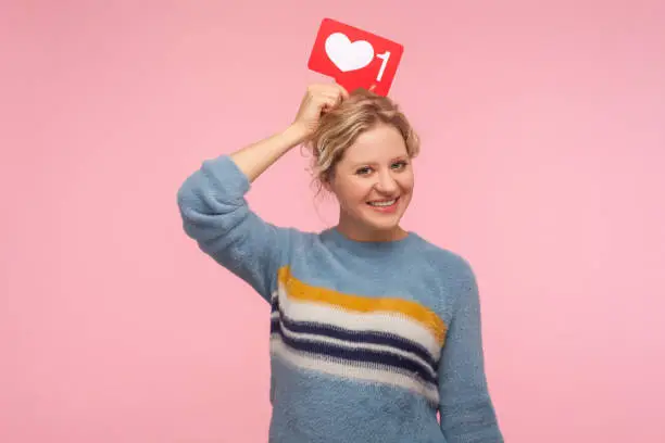Photo of Portrait of cheerful adult woman with curly hair in warm sweater holding heart Like icon over head, love content